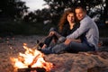 Night camping on shore. Happy man and woman hikers having a rest in front of tent at campfire and forest background