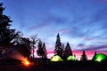 Night camping. Tourist have a rest at a campfire near illuminated tent and wooden house under amazing night sky full of