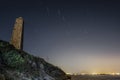 Night image of a beach with a stone monolith, lights and city in the background and a line of a star forming a circumpolar, in