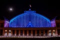 Night photo of the facade of Atocha station illuminated with colored lights and the moon in the background