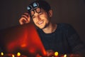 Night portrait of young happy man wearing round eyeglasses, looking in laptop in dark room with garlands at home. Royalty Free Stock Photo