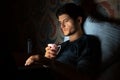 Night portrait of young man lying on bed in dark room at home with cup of coffee and laptop on legs.