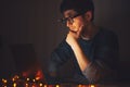 Night portrait of young thoughtful man wearing round glasses, looking in laptop in dark room with garlands at home. Royalty Free Stock Photo