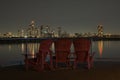 Night scene of wooden chairs at the waterfront lookout with a view of night lights of a cityscape.