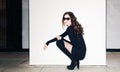 Night shot fashion portrait of brunette model in summer black clotches and sunglasses sitting near white wall.