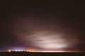 Night Sky With Rare Glowing Stars Shining Through The Cloudiness Overcast Above Countryside Town Landscape. Rural Field