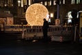 Night street scene in Berlin city. Huge shiny golden ball in darkness and human silhouette near road construction striped fencing