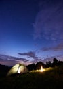 Burning bonfire near the tent in which light is on against backdrop of powerful mountains under starry sky