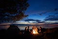 Night summer camping on shore. Group of young tourists around campfire near tent under evening sky