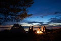 Night summer camping on shore. Group of young tourists around campfire near tent under evening sky Royalty Free Stock Photo
