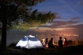 Night summer camping on shore. Group of young tourists around campfire near tent under evening sky Royalty Free Stock Photo