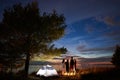 Night summer camping on shore. Group of young tourists around campfire near tent under evening sky Royalty Free Stock Photo