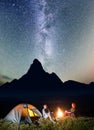 Night tent camping. Tourist couple - girl and guy sitting by bonfire under incredibly beautiful starry sky