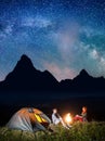 Night tent camping. Young couple backpackers sitting by bonfire under incredibly beautiful starry sky and Milky way