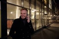 Night time, one young smiling and happy man, 20-29 years, talking over his phone, standing in Autumn coat on street, in front of Royalty Free Stock Photo