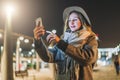 Night. Young woman is standing on city street and is looking at screen of smartphone. Girl uses digital gadget, chatting Royalty Free Stock Photo