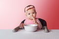 Nine-month-old baby girl in pink bandage sits at white table in highchair, eats herself with spoon from bowl. Pink background Royalty Free Stock Photo