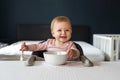 Nine-month-old smiling baby girl sits at white table in highchair and eats herself with spoon from bowl. Blurred background Royalty Free Stock Photo