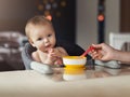 Nine-month-old smiling baby girl sits at white table in highchair, eats with spoon from bowl. Mom feeds baby from spoon