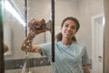 Nobody does it better. Young smiling afro american woman in uniform cleaning shower door with squeegee and spray