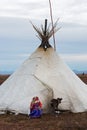 Ousewife of nenets nomad tribe in national costume repairs a clothes in front of the family chum with young reindeer.