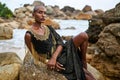 Non-binary black person in luxury dress on rocks in ocean. Trans ethnic fashion model wearing jewellery dressed in posh