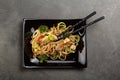 Noodles with meat and vegetables in black plate, chopsticks, on grey surface Royalty Free Stock Photo