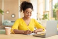 Note taking concept. Happy young black woman using pc laptop and writing in her workbook, sitting at desk at home Royalty Free Stock Photo