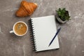 Notebook, cup of aromatic coffee, delicious croissant and plant on brown marble table, flat lay