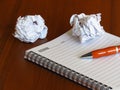 A notebook, an orange pen and two smashed paper sheet balls on dark wood background.