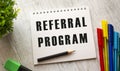 A notebook on a spring with the text REFERRAL PROGRAM lies on a wooden table with colored pens Royalty Free Stock Photo