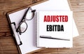 Notebook with text adjusted EBITDA on wooden desk with glasses and clipboard Royalty Free Stock Photo