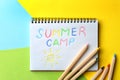 Notebook with written text SUMMER CAMP and different pencils on color background
