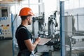 With notepad in hands. Industrial worker indoors in factory. Young technician with orange hard hat