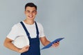 With notepad and hard hat. Male worker in blue uniform standing inside of studio against white background Royalty Free Stock Photo