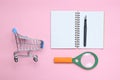 Notepad, magnifier, shopping trolley on pink patel background.