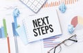 Notepad with tex NEXT STEPS on the business charts and pen,business Royalty Free Stock Photo