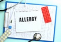 A notepad with the text ALLERGY lies on a medical clipboard with a stethoscope and pills on a blue background. Royalty Free Stock Photo