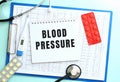 A notepad with the text BLOOD PRESSURE lies on a medical clipboard with a stethoscope and pills on a blue background. Royalty Free Stock Photo