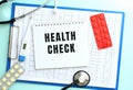 A notepad with the text HEALTH CHECK lies on a medical clipboard with a stethoscope and pills on a blue background. Royalty Free Stock Photo