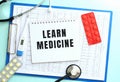 A notepad with the text LEARN MEDICINE lies on a medical clipboard with a stethoscope and pills on a blue background. Royalty Free Stock Photo