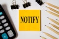 Notify word written on a yellow piece of paper on a white background with office supplies lying nearby