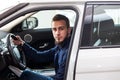 A young dark-haired guy with a beard sits behind the wheel of a white car and looks into the camera through the open door with one