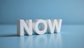 NOW word in white block letters on blue surface with soft shadow and reflection, conveying urgency and focus in calm setting Royalty Free Stock Photo