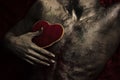 Nude torso with red plush soft heart toy, dark background.