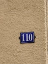 Number 110 house number plate. Blue plate with white numbers on a pale yellow building wall, no people