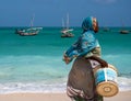 NUNGWI, ZANZIBAR - JAN 20, 2019: local woman in Bright Cloth walking by the Indian ocean Beach with a Bucket for Fish