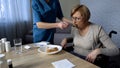 Nurse helping sick old female to eat, medical support and care, sadness