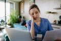 Nurse paying bills online, sitting in living room, working on notebook. Doctor in scrubs dealing with work issues from Royalty Free Stock Photo