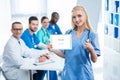 happy nurse holding tablet and showing thumb up with colleagues sitting at table